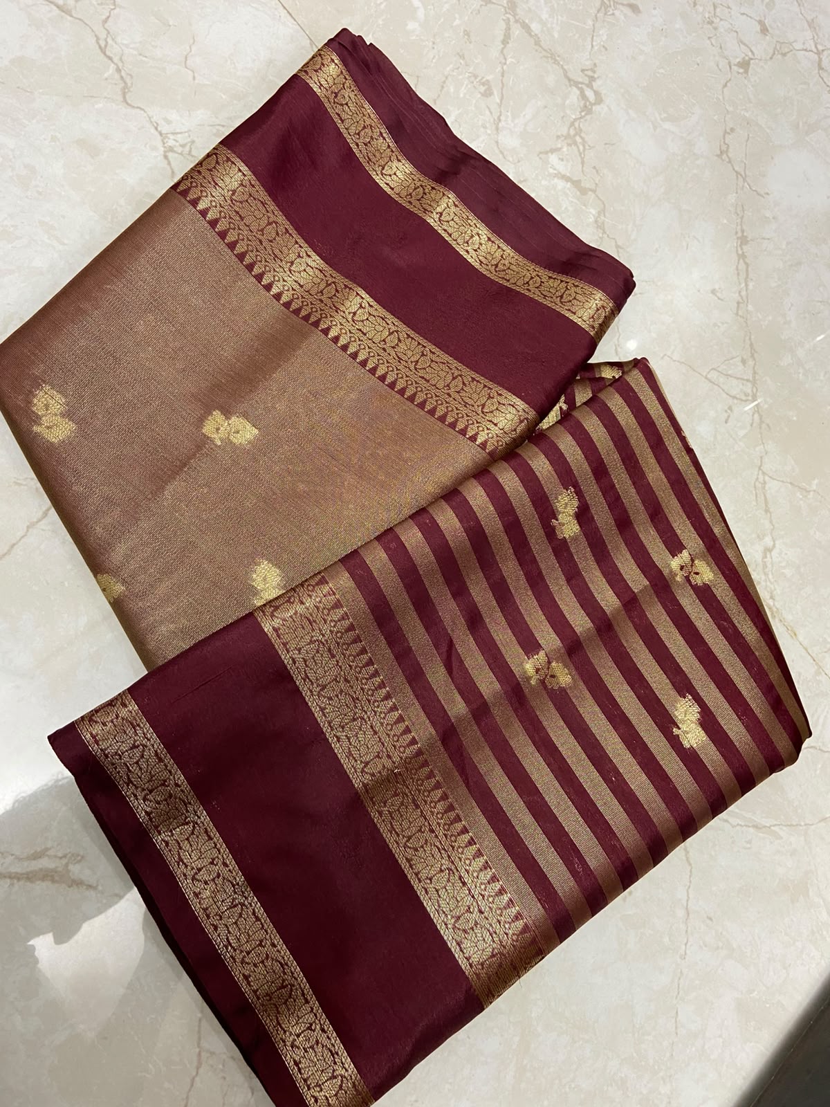 A folded silk saree displayed on a light beige marble surface, featuring a deep maroon border, a beige body with small gold motifs, and alternating maroon and gold stripes with a decorative gold zari border.