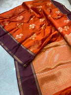 An orange and deep purple silk saree with intricate gold zari floral motifs and a detailed gold-embellished border, laid out on a light marble surface.