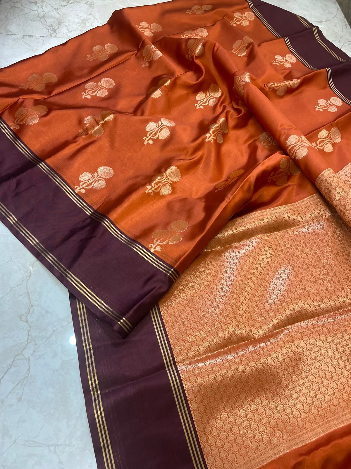 An orange and deep purple silk saree with intricate gold zari floral motifs and a detailed gold-embellished border, laid out on a light marble surface.