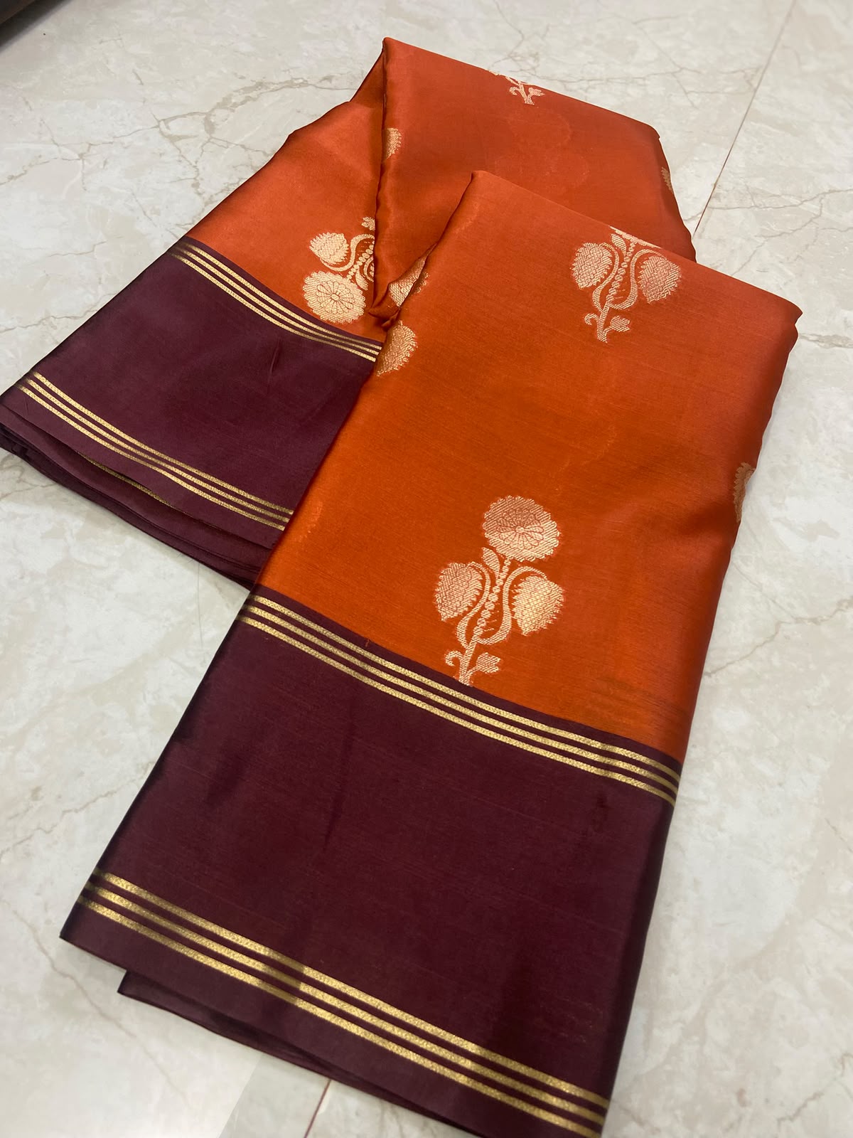 An orange and deep maroon silk saree with gold zari floral motifs and borders, folded on a light marble floor.