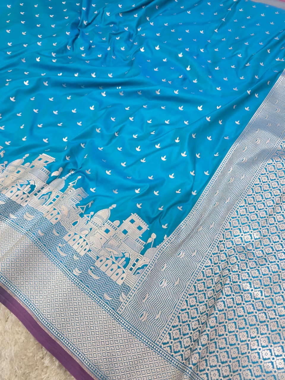 A blue and silver Banarasi silk saree with white zari embroidery, featuring a cityscape motif on the border and a geometric pattern on the pallu.
