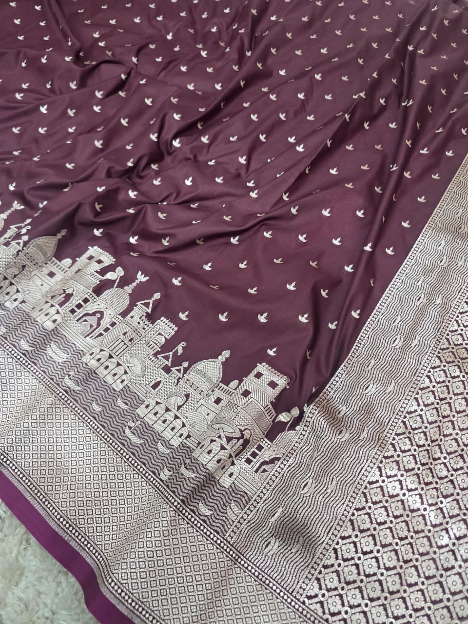 A close-up of a purple fabric, likely a saree, featuring a silver-toned border with architectural motifs and a scattered pattern of small bird silhouettes.