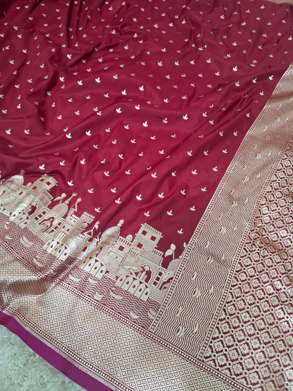 A deep red Banarasi silk saree with intricate gold zari embroidery, featuring a detailed border with palace motifs and a geometric pattern.