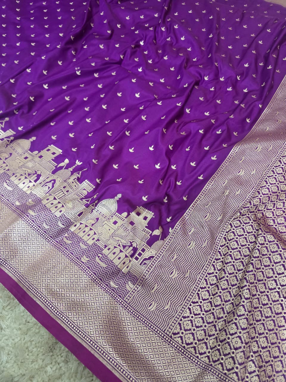 A purple silk saree with intricate silver zari embroidery, featuring a border with a cityscape motif and geometric patterns.