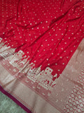 A close-up of a red fabric, likely a saree or scarf, featuring small white motifs across the body and a wide beige border with a detailed architectural pattern.