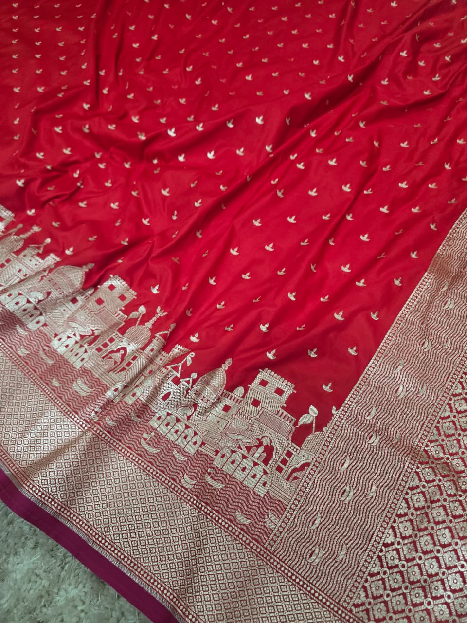 A close-up of a red fabric, likely a saree or scarf, featuring small white motifs across the body and a wide beige border with a detailed architectural pattern.