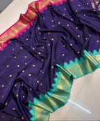 A dark purple silk saree with gold zari motifs, a vibrant pink border, and a green and gold zari pallu, draped on a light gray tiled floor.
