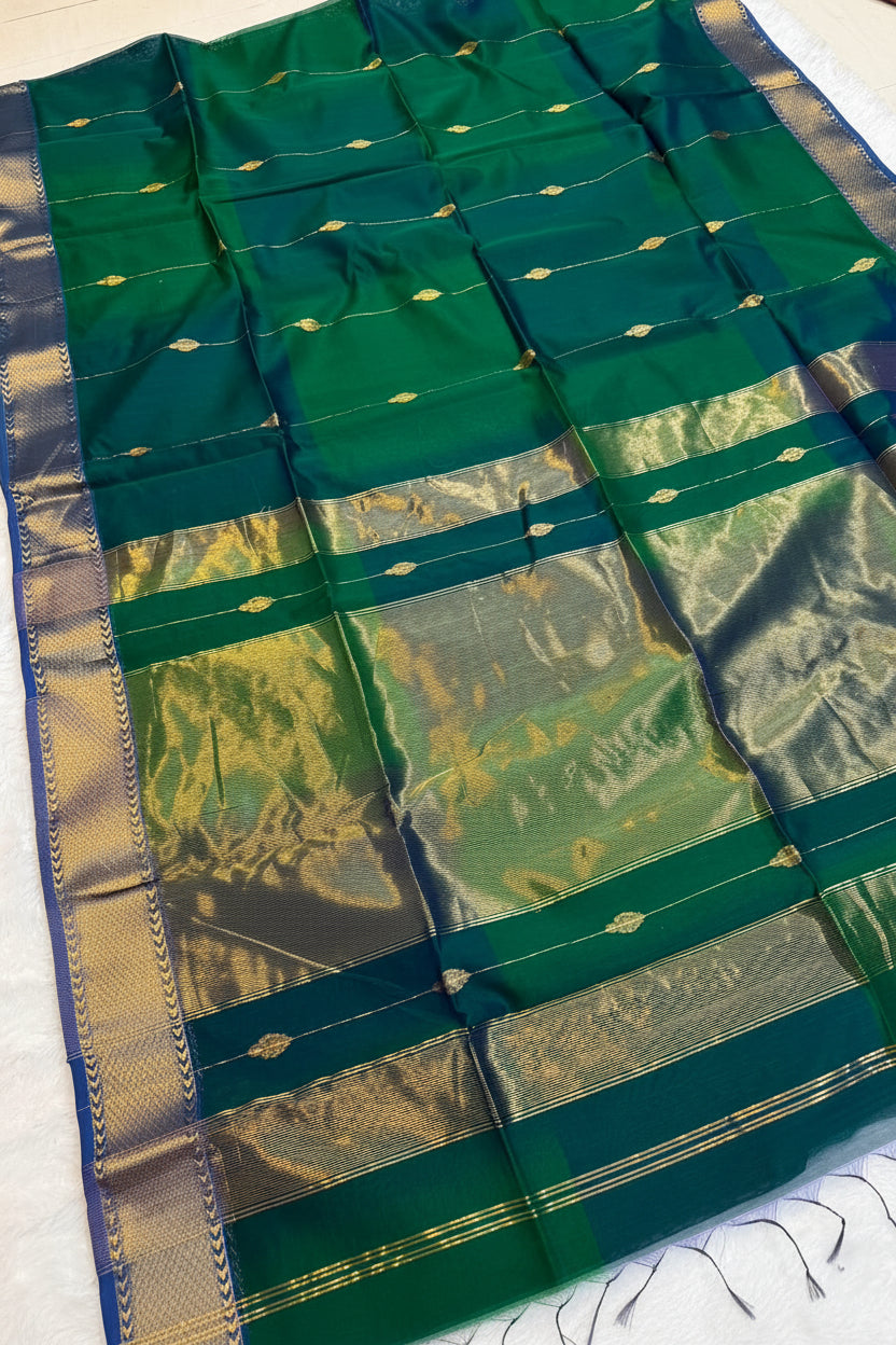 A dark green silk saree with a wide gold zari border and metallic gold motifs, laid flat on a white surface with a small decorative flower arrangement.