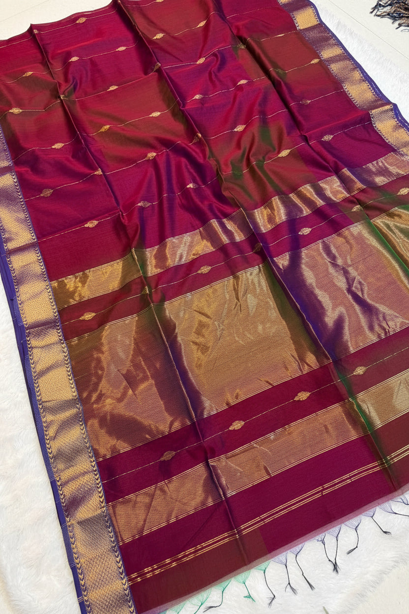 A deep magenta-purple silk saree with a wide gold and beige zari border, laid out diagonally on a white surface with a small decorative flower basket.