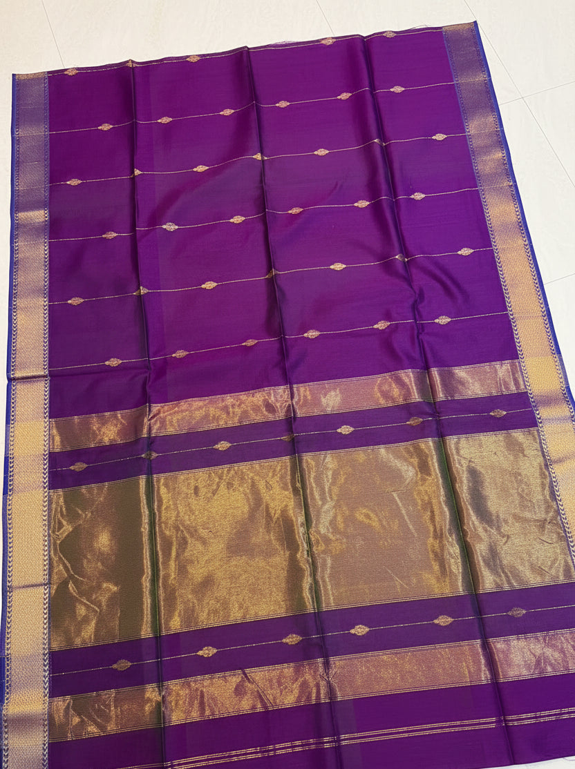 A purple silk saree with metallic gold zari stripes and motifs, laid out on a light-colored floor with a decorative basket of flowers on top.