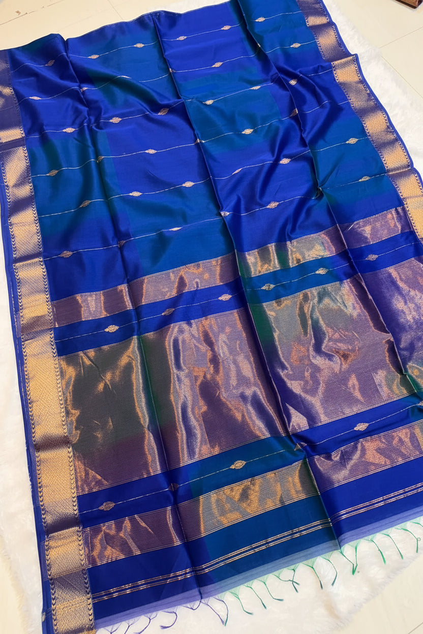 A royal blue silk saree with metallic gold and silver zari borders, laid flat on a white surface with a decorative basket of flowers on the border.