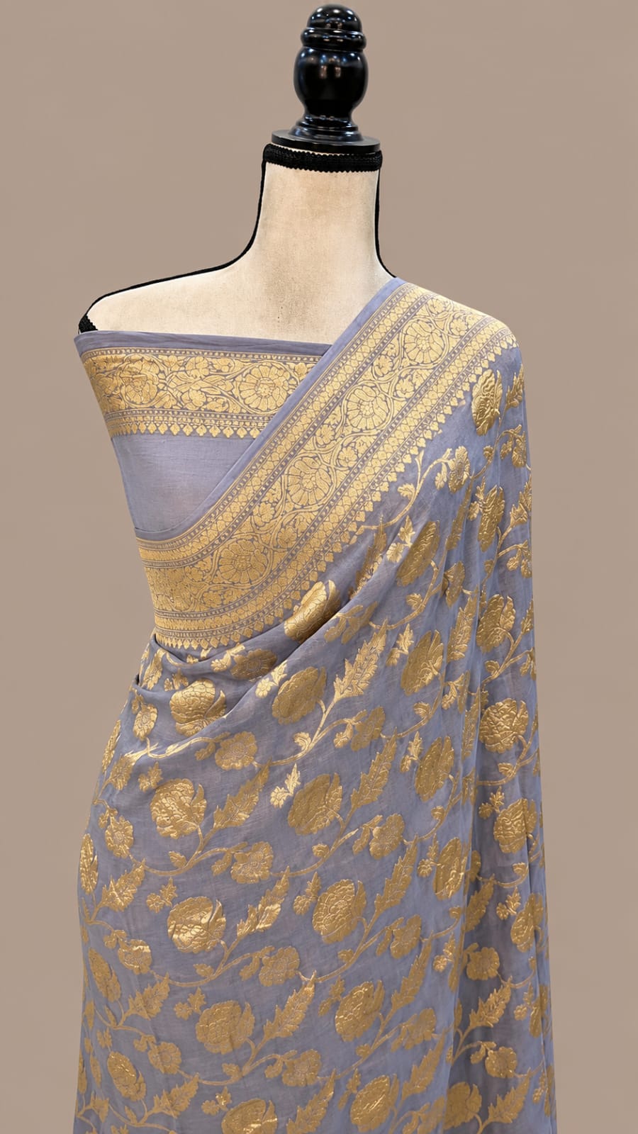 A lavender blue saree with intricate gold floral zari embroidery, draped on a mannequin torso against a solid beige background.