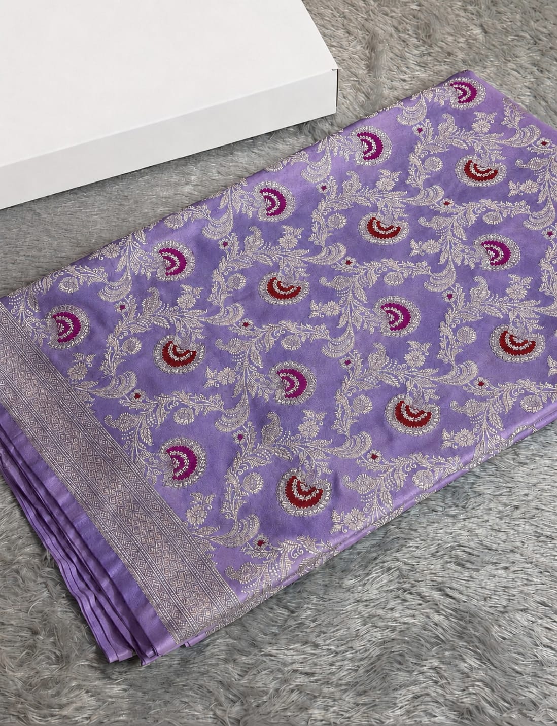 A folded purple silk saree with intricate silver floral patterns and red paisley motifs, displayed on a gray faux fur surface next to a white box.