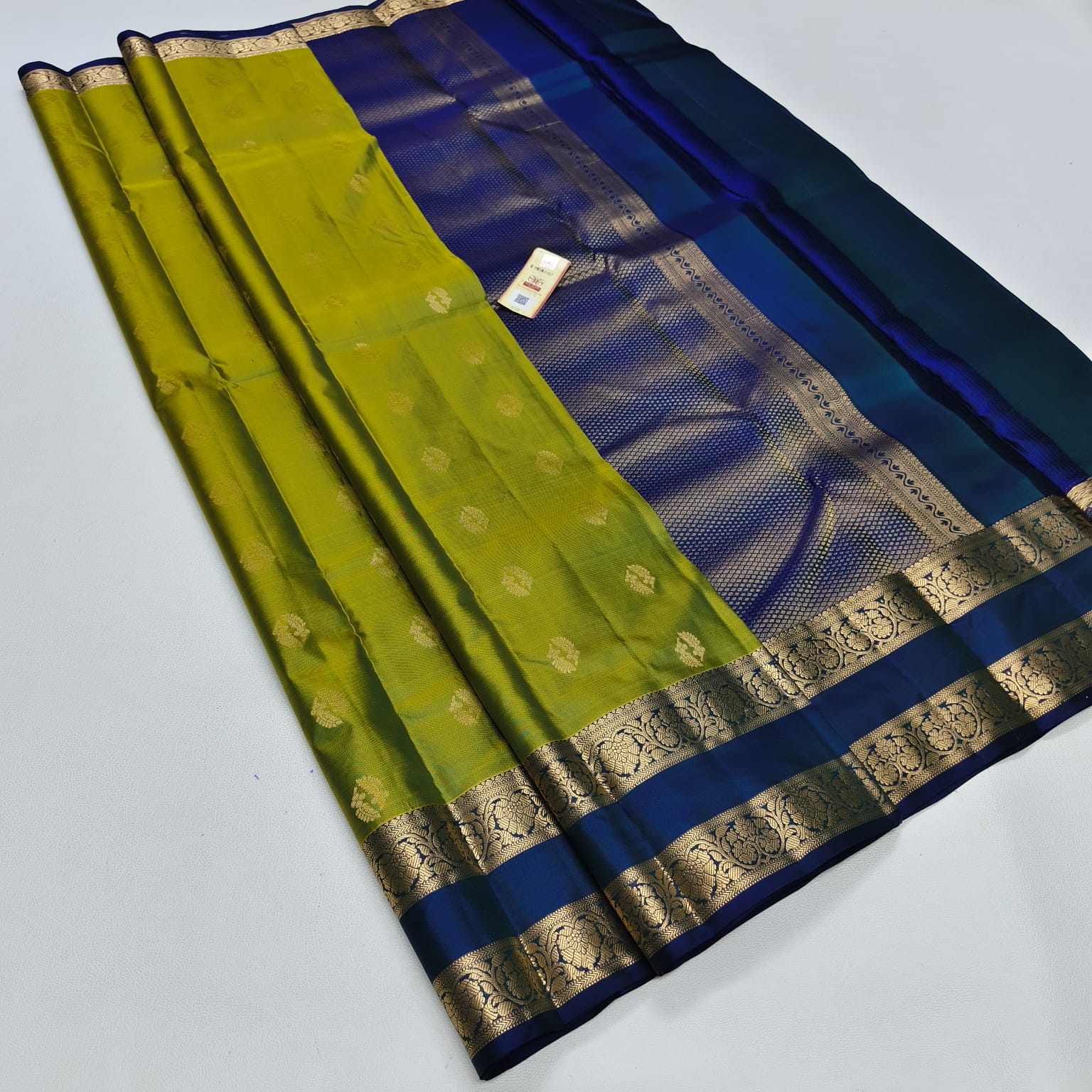 A folded silk saree with a vibrant olive green body, a rich royal blue border, and intricate gold zari woven patterns, displayed on a light gray background.