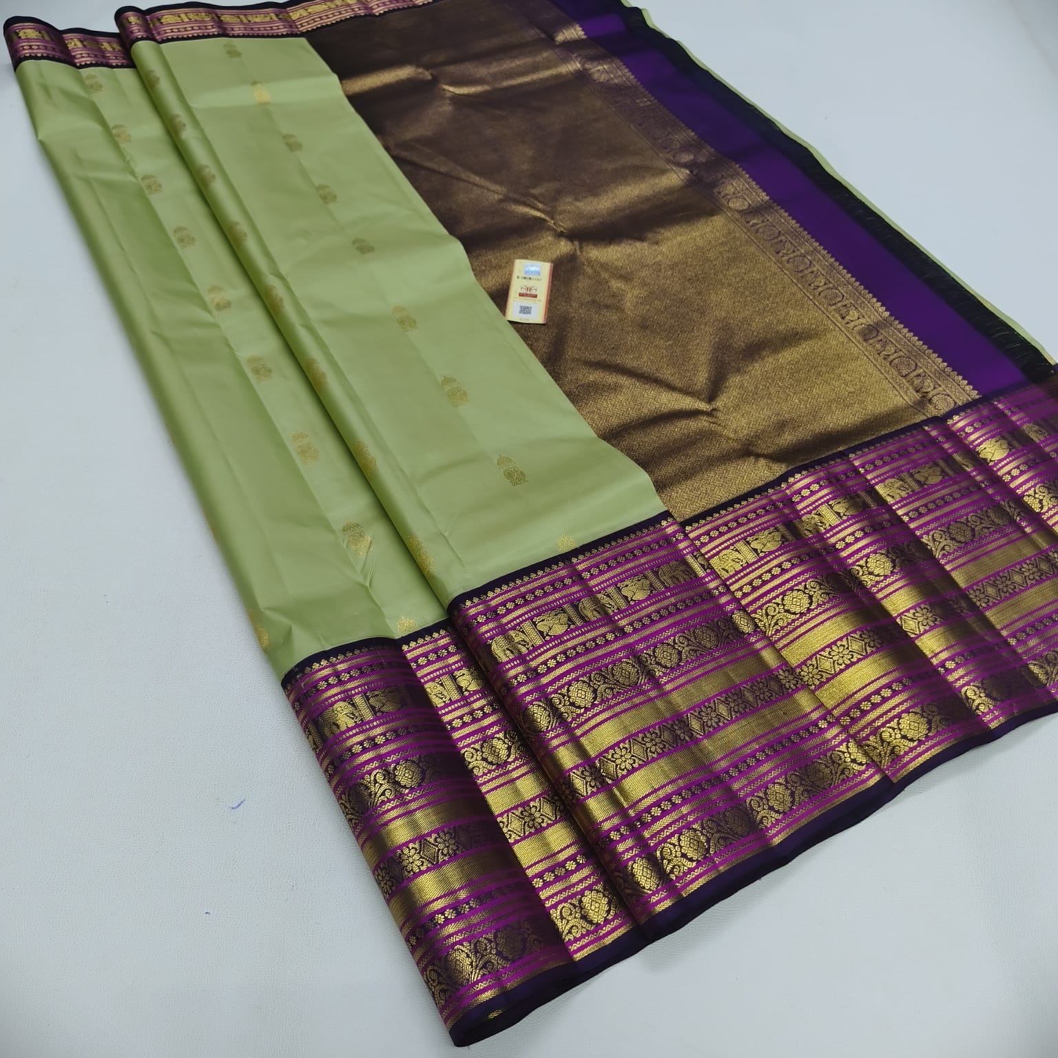 A traditional Indian silk saree with a light olive green body, a metallic gold and brown zari-pallu, and a wide purple and gold brocade border.