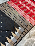 A folded and draped saree fabric with a black body, a red border, and intricate metallic gold and silver zari embroidery, laid out on a gray marble surface.