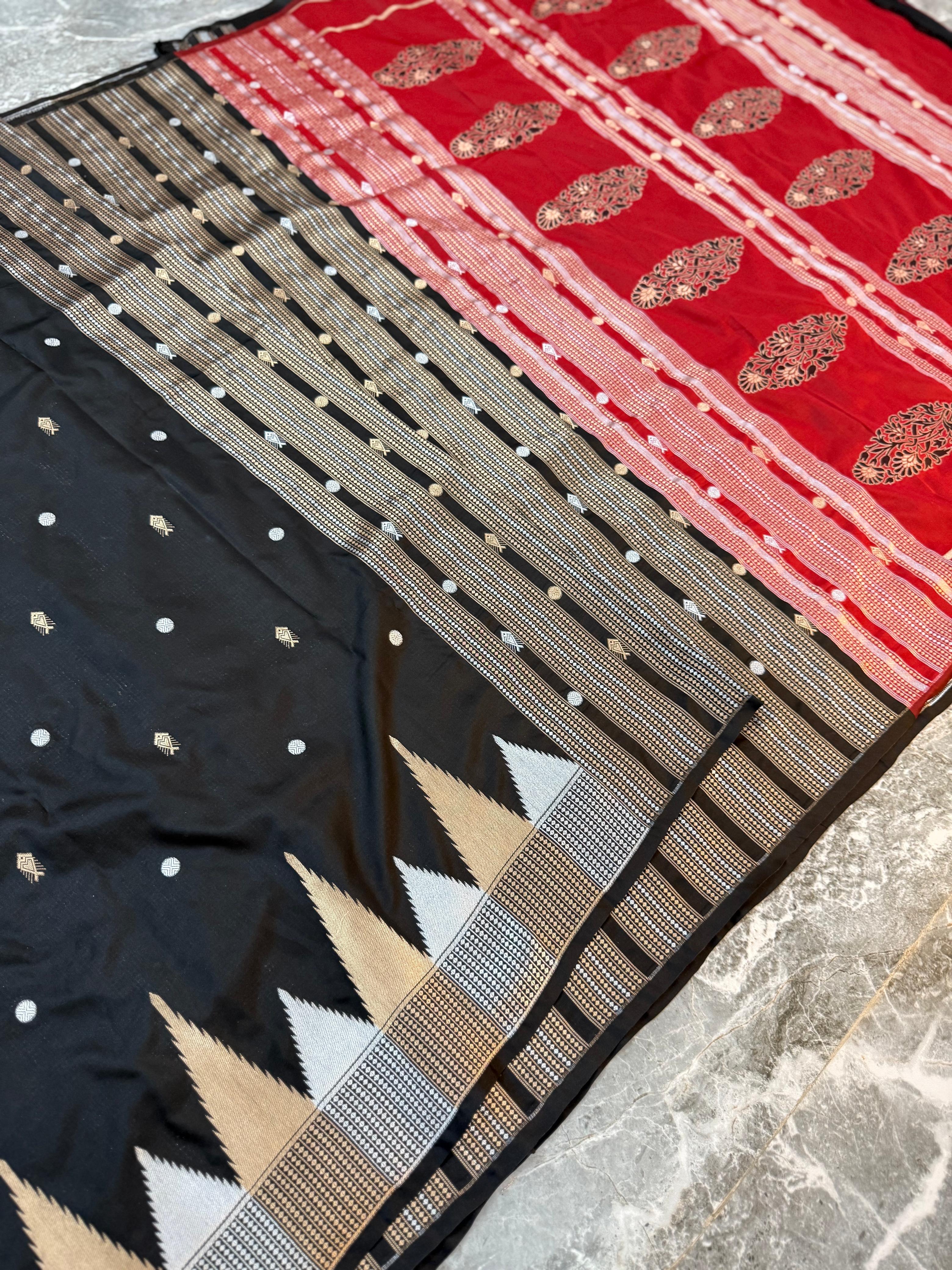 A folded and draped saree fabric with a black body, a red border, and intricate metallic gold and silver zari embroidery, laid out on a gray marble surface.