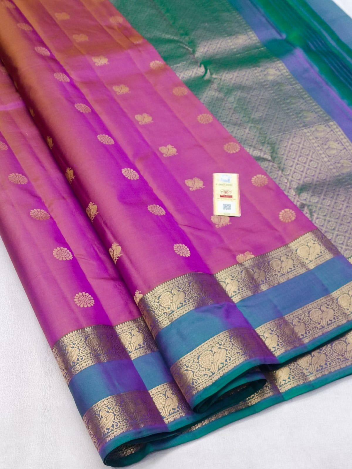 A folded silk saree with a vibrant magenta body, a teal green border, and intricate gold zari woven patterns, displayed on a light gray surface.