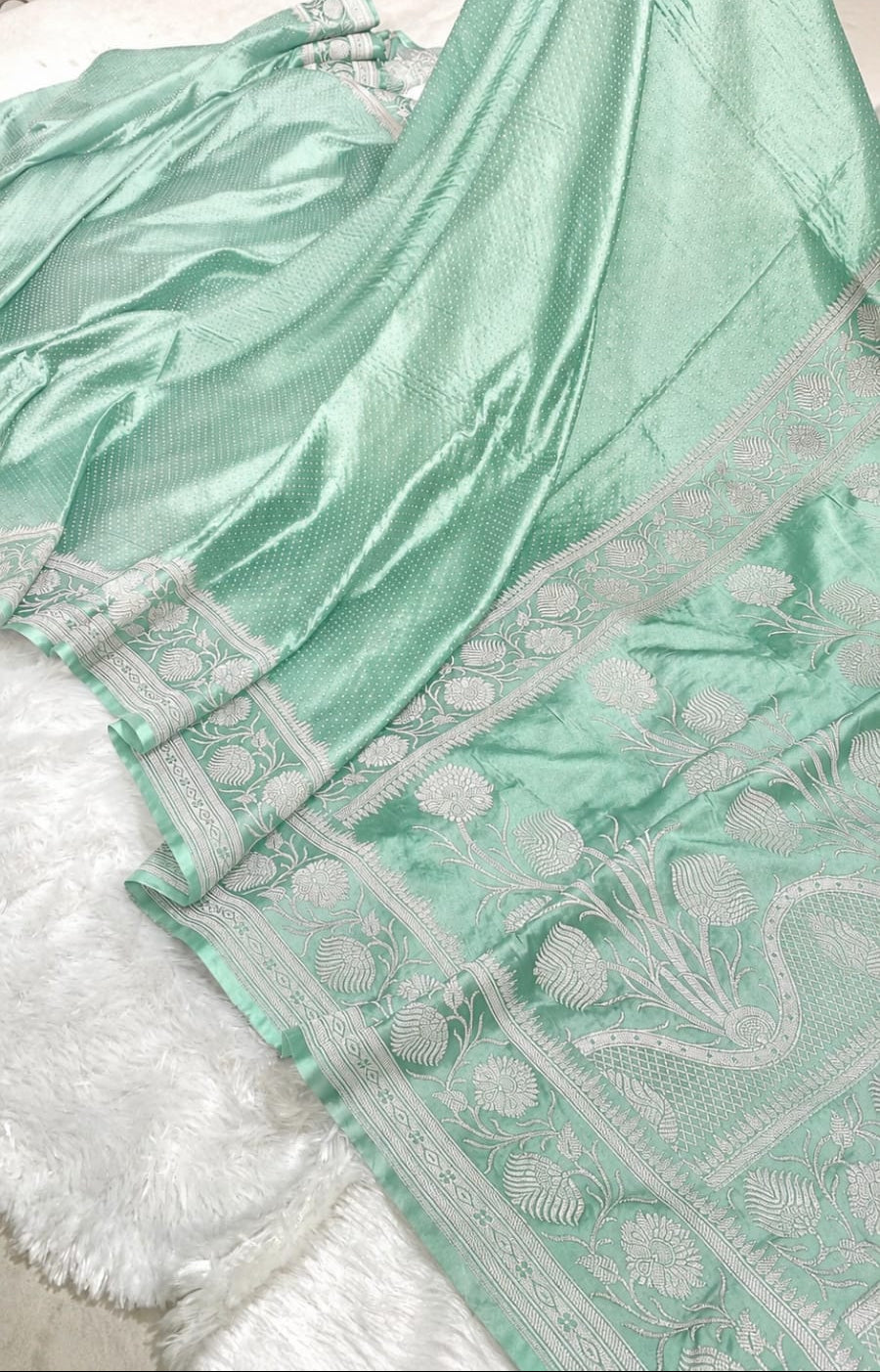 A mint green silk saree with a wide white border featuring floral and geometric patterns, draped over a white faux fur surface.