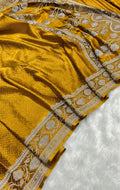 A close-up of a mustard yellow silk saree with intricate silver and white floral embroidery along its border, laid out on a white textured surface.