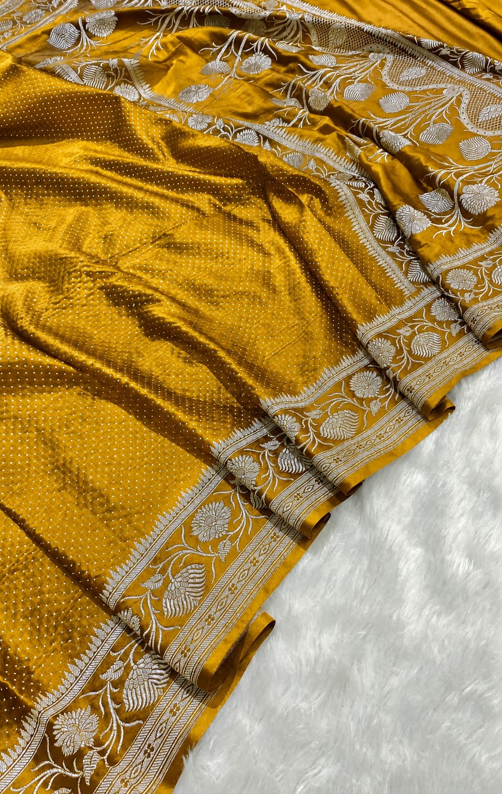 A close-up of a mustard yellow silk saree with intricate silver and white floral embroidery along its border, laid out on a white textured surface.