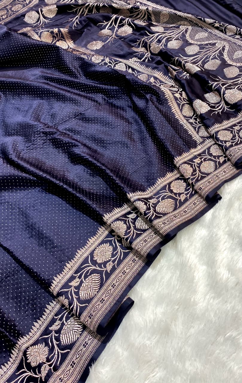 A close-up of a dark navy blue fabric, likely a saree or scarf, featuring a textured body with tiny dot patterns and a wide, ornate border decorated with beige and gold floral motifs.