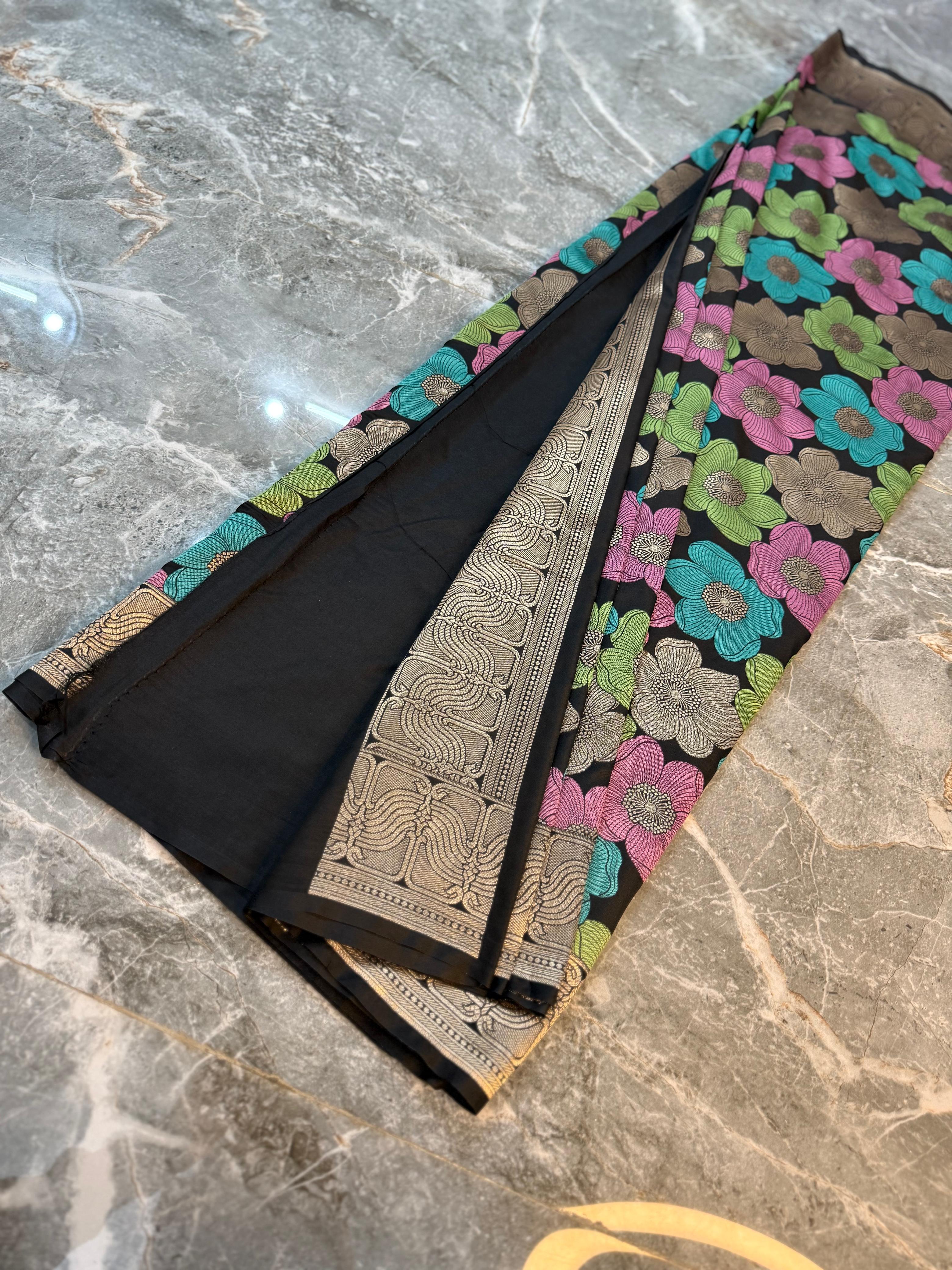 A folded black fabric garment, likely a saree or dupatta, displayed on a gray marble surface. The fabric features a prominent floral pattern in pink, green, and blue, with a wide beige and black patterned border.
