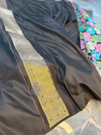 A close-up of a black fabric garment, likely a saree, featuring a wide metallic silver border with gold and silver floral patterns and a yellow-gold section. A portion of a colorful floral fabric is visible in the background.