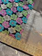 A close-up of a richly decorated fabric, likely a saree, featuring a navy blue base with vibrant floral patterns in pink, teal, green, and silver. The fabric is adorned with intricate metallic borders in gold and silver, showcasing detailed woven textures.