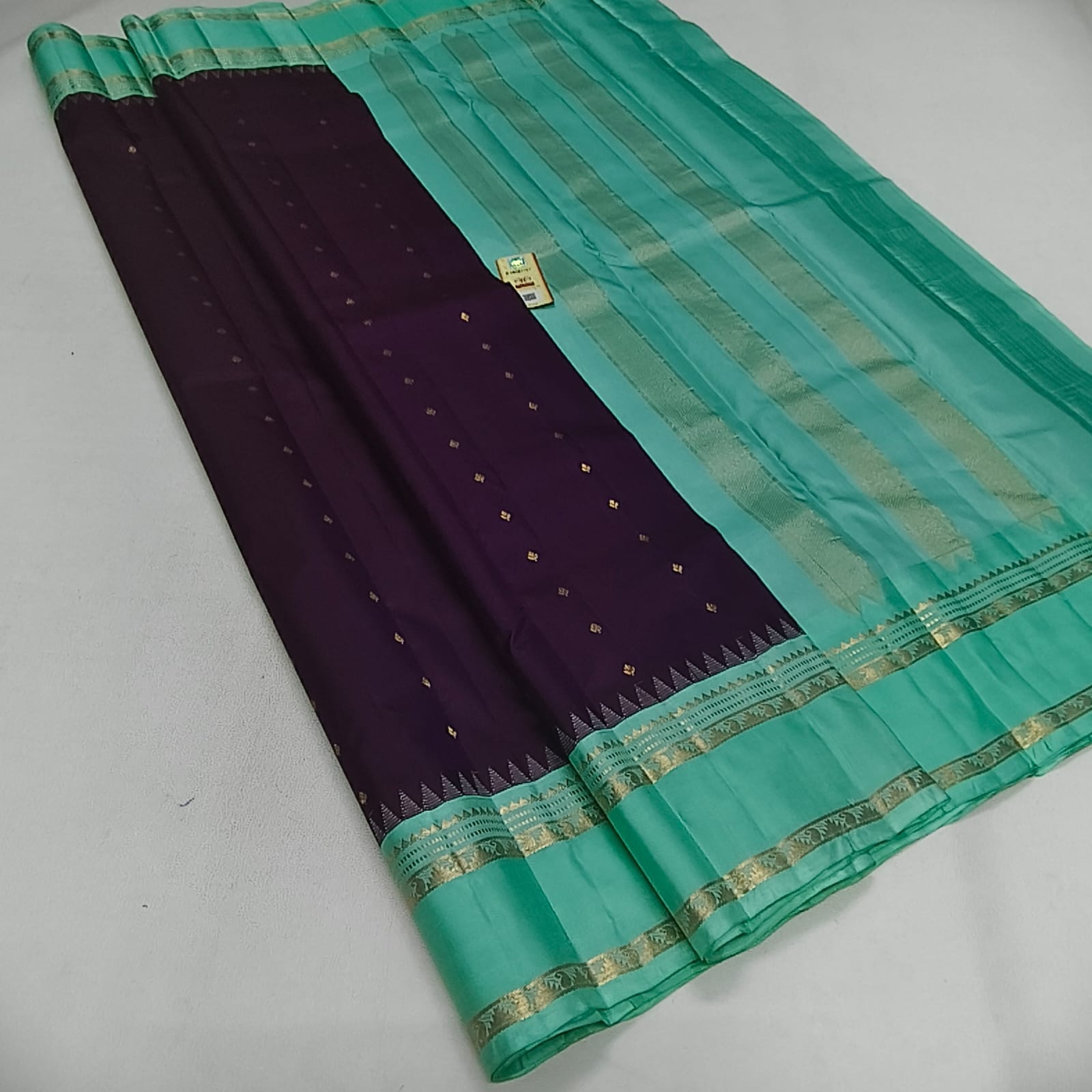 A folded silk saree with a deep purple body, decorated with small gold zari motifs, and a vibrant teal-green border with metallic gold stripes.
