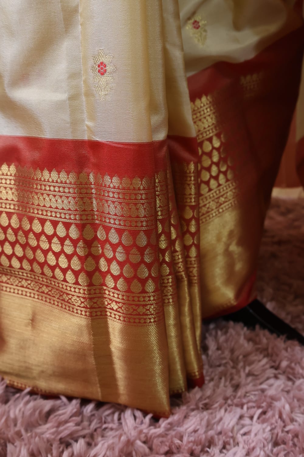 A close-up of a cream-colored silk saree with a rich red border and intricate gold zari woven patterns, draped over a pink shag rug.