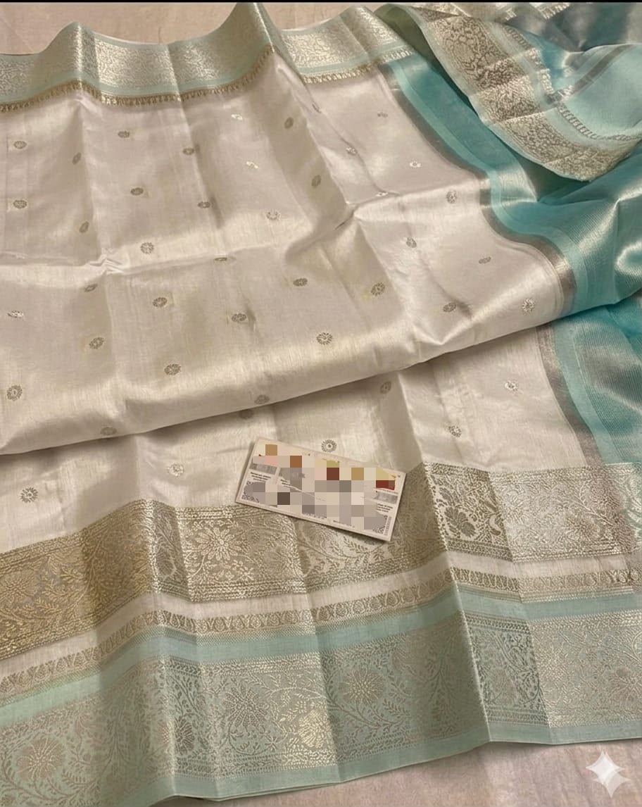 A close-up of a light-colored silk or satin fabric with metallic gold woven borders and small gold dot motifs, accented with a soft mint green border. A small card is placed on the fabric.
