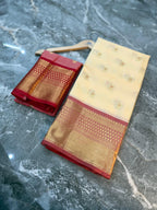 A folded cream-colored silk saree with a red border and gold zari motifs, displayed alongside its matching red and gold pleated fabric cover on a gray marble surface.