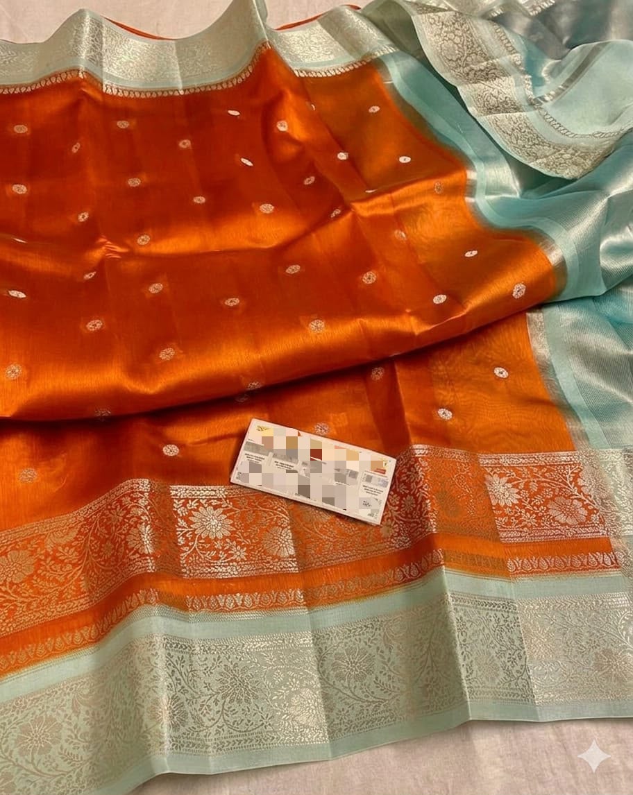 An orange silk saree with a light blue border and silver zari brocade work, featuring small metallic motifs throughout. A small card is placed on the fabric.