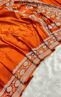 A close-up of an orange silk saree with intricate silver and white floral border patterns, draped over a white textured surface.