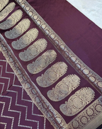 A close-up of a deep purple fabric, likely a saree, featuring a wide border with intricate gold zari embroidery in paisley motifs and a zigzag patterned section.