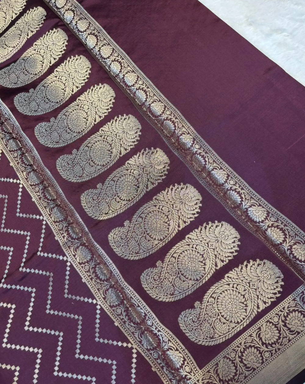 A close-up of a deep purple fabric, likely a saree, featuring a wide border with intricate gold zari embroidery in paisley motifs and a zigzag patterned section.