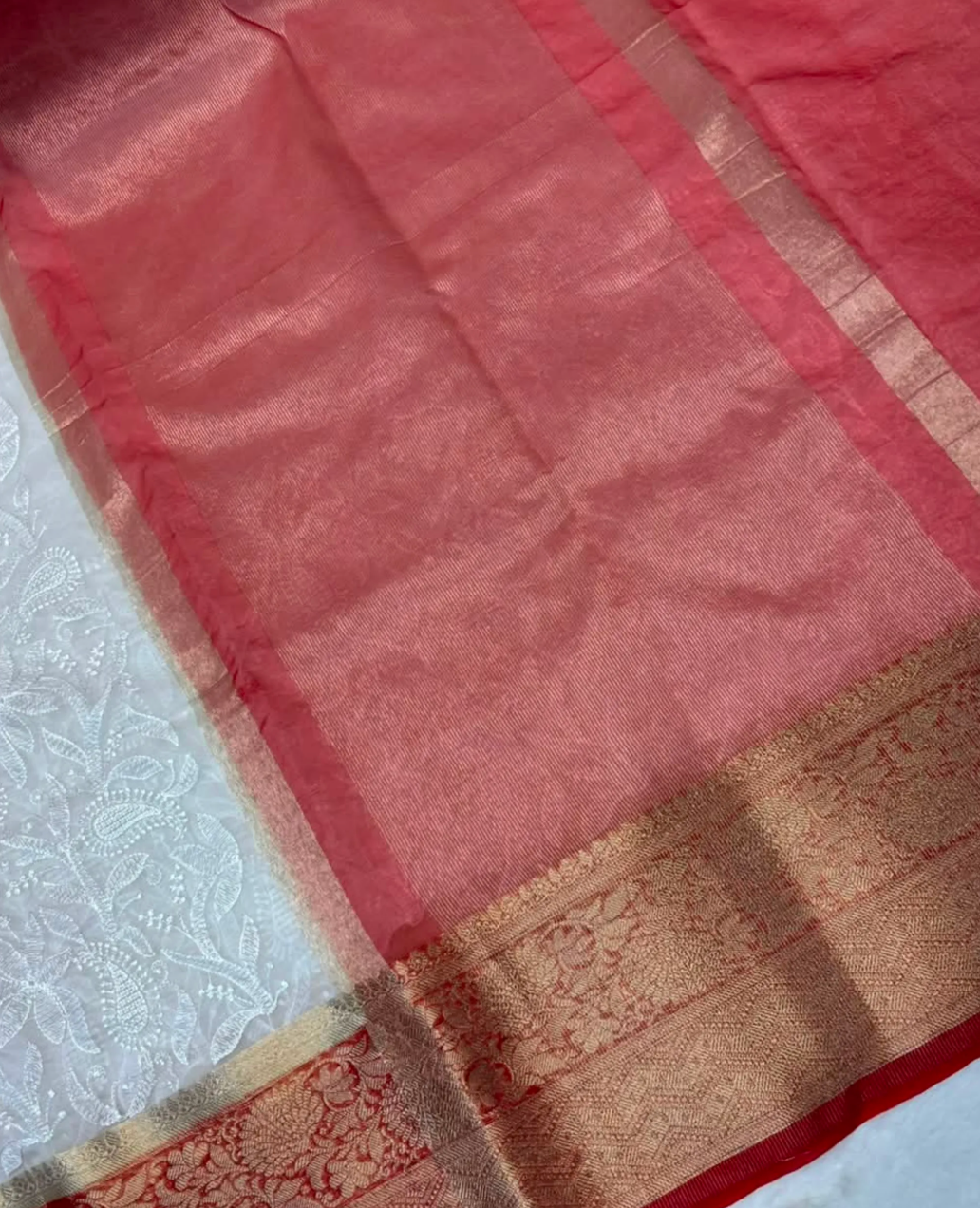A close-up of a pink silk saree body with a wide gold and beige zari border, laid flat next to a white embroidered fabric.