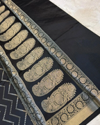 A close-up of a black fabric textile with ornate metallic gold woven paisley and floral border patterns, laid diagonally on a light neutral surface.