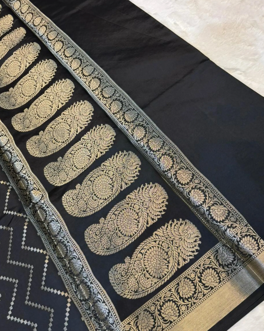 A close-up of a black fabric textile with ornate metallic gold woven paisley and floral border patterns, laid diagonally on a light neutral surface.