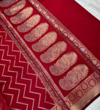 A close-up of a rich red fabric, likely a saree, featuring intricate gold zari woven patterns including paisley motifs and a zigzag border.