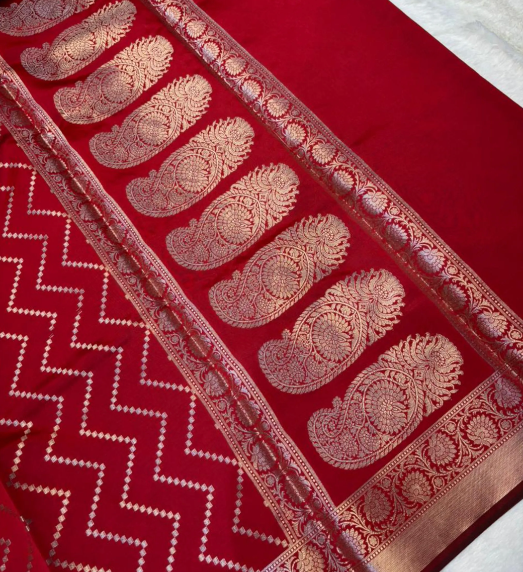 A close-up of a rich red fabric, likely a saree, featuring intricate gold zari woven patterns including paisley motifs and a zigzag border.