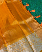 A close-up of a golden-yellow silk saree or fabric with a silver zari border and orange tassels, laid over a textured green surface.