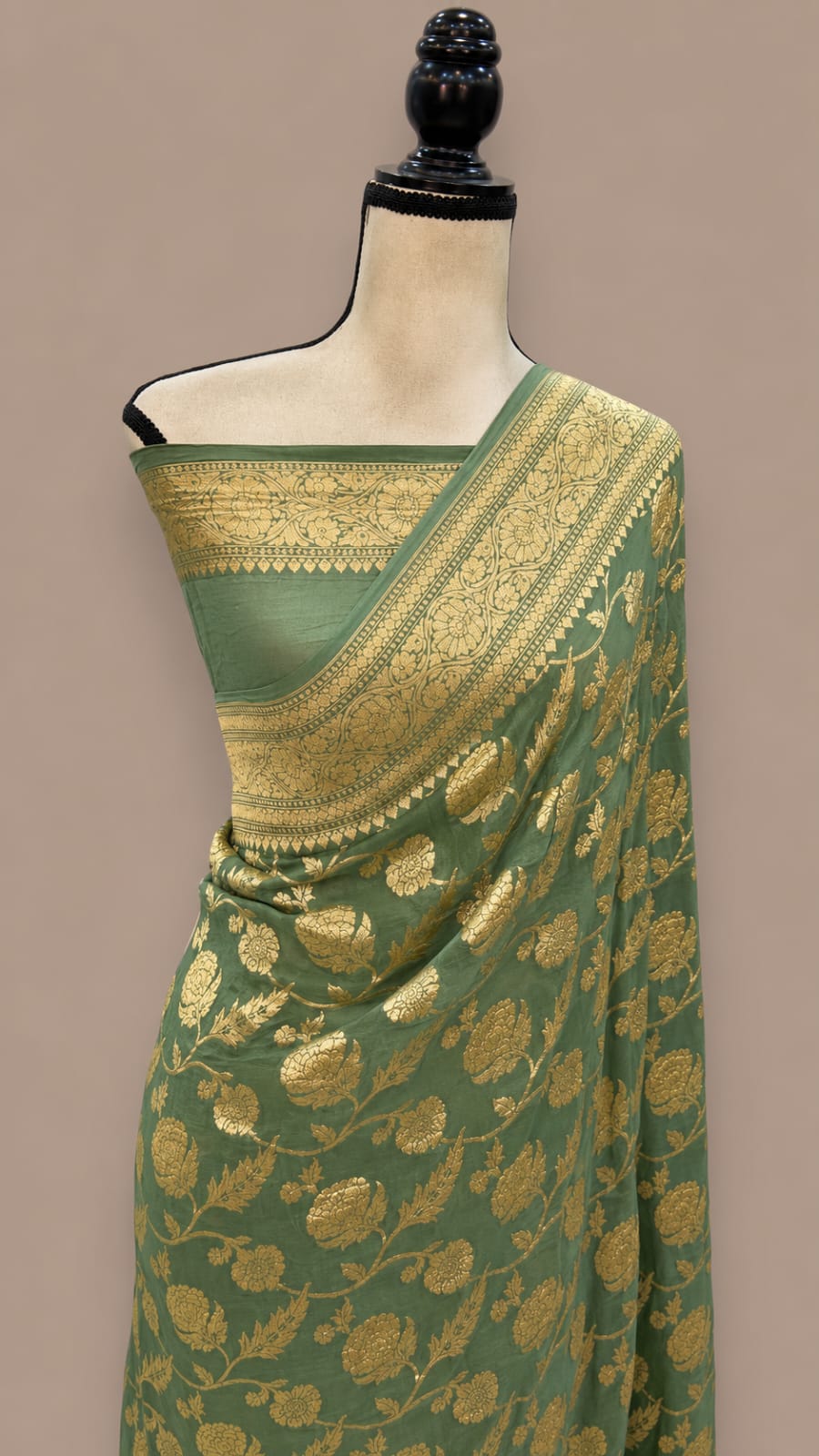 A sage green silk saree with intricate gold zari floral motifs and a wide decorative gold border, displayed on a cream-colored mannequin torso against a solid taupe background.
