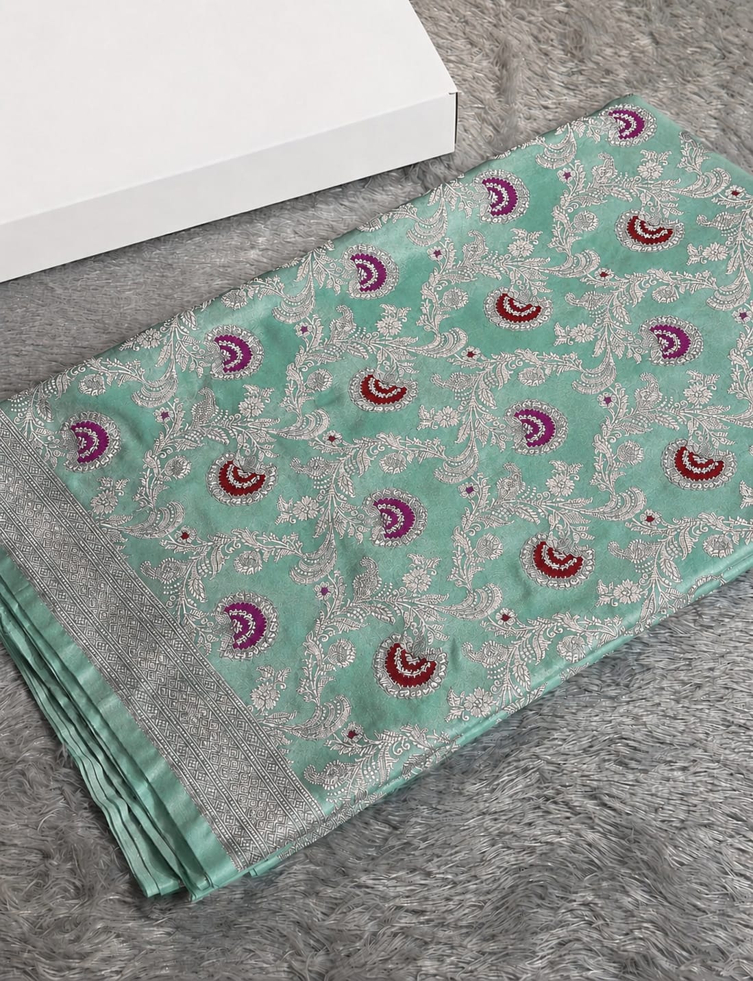 A folded mint green silk saree with intricate silver zari embroidery and purple/magenta paisley motifs, displayed on a gray faux fur surface next to a white box.