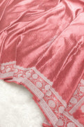 A close-up of a pink fabric with a textured dotted pattern and a white floral brocade border, laid over a white faux fur surface.