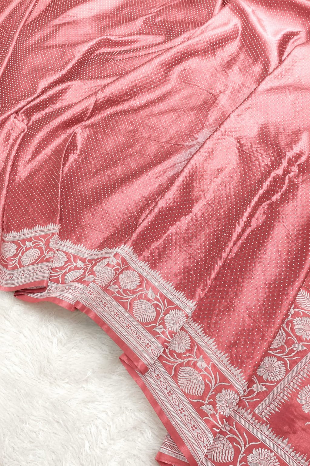 A close-up of a pink fabric with a textured dotted pattern and a white floral brocade border, laid over a white faux fur surface.