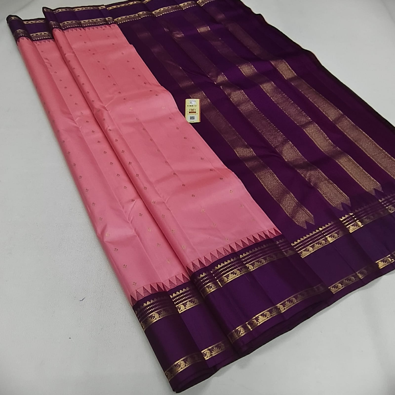 A traditional Indian saree displayed diagonally on a light gray surface, featuring a pink body with subtle gold motifs, a deep purple border, and gold zari metallic stripes.