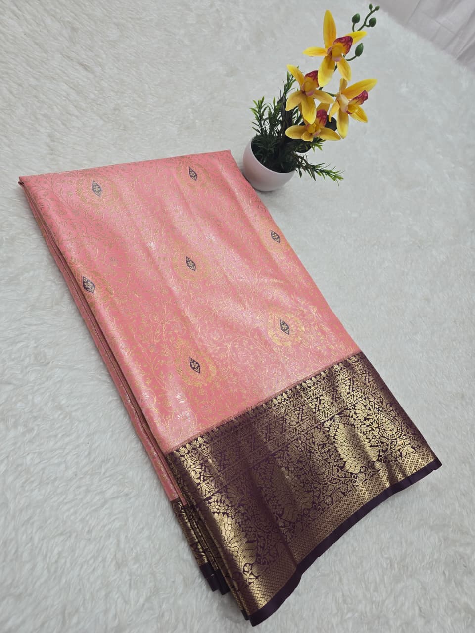 Pink saree with a brown and gold border on a light gray surface with a small plant.