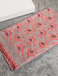 A folded coral pink saree with intricate silver floral embroidery and red and purple paisley motifs, displayed on a gray textured rug next to a white box.